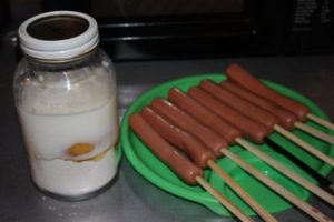 Photo of a hot dogs on chopsticks and a jar of milk and eggs for an egg wash to deep fry.