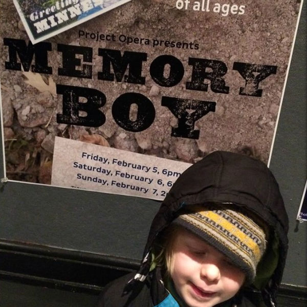Young boy standing in front of poster that reads "Project Opera presents Memory Boy. Opera for kids of all ages."