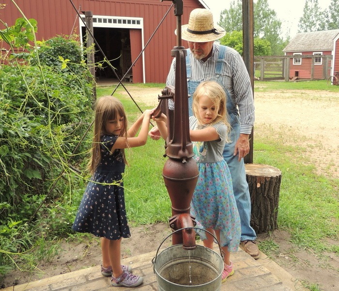 Costumed actor helping two girls to use water pump at Eidem Homestead in Brooklyn Park, MN