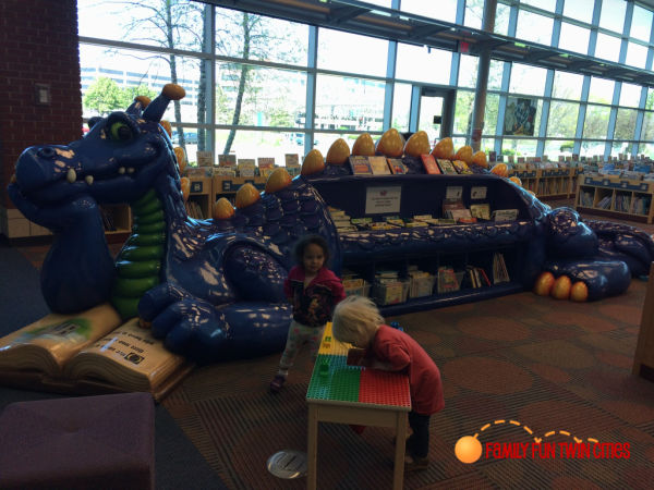 Little girls exploring the children's section of Brookdale Library in A bookshelf in the shape of a purple dragon reading a book lounges in the background. Summer Reading Programs at Twin Cities Libraries