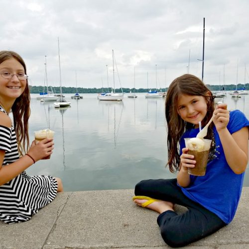 Two girls watching sailboats as they eat Rootbeer Floats from The Bread & Pickle at Lake Harriet Park in Minneapolis, Minnesota