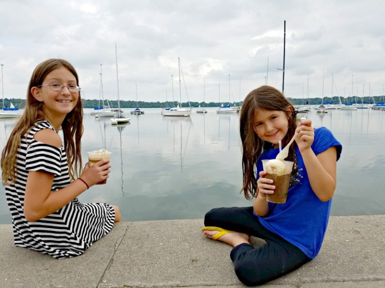 Two girls watching sailboats as they eat Rootbeer Floats from The Bread & Pickle at Lake Harriet Park in Minneapolis, Minnesota