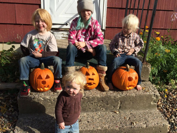 Three kids showing off their carved pumpkins as a toddler looks on