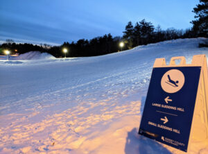 Sledding hill at Battle Creek Park lit up at night. Sign pointing to large sledding hill and small sledding hill.