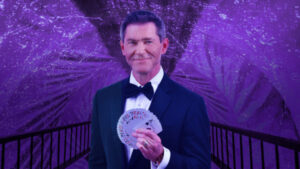 man in tuxedo holding playing cars with a purple background